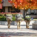 Three ROTC students carrying large packs walk through the Orange Grove on an afternoon in Autumn.